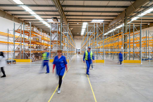 Two workers in safety vests walking through a wide warehouse aisle flanked by tall metal shelving units, illustrating the scale of commercial storage operations and the importance of fire code compliance for lithium battery storage container areas.