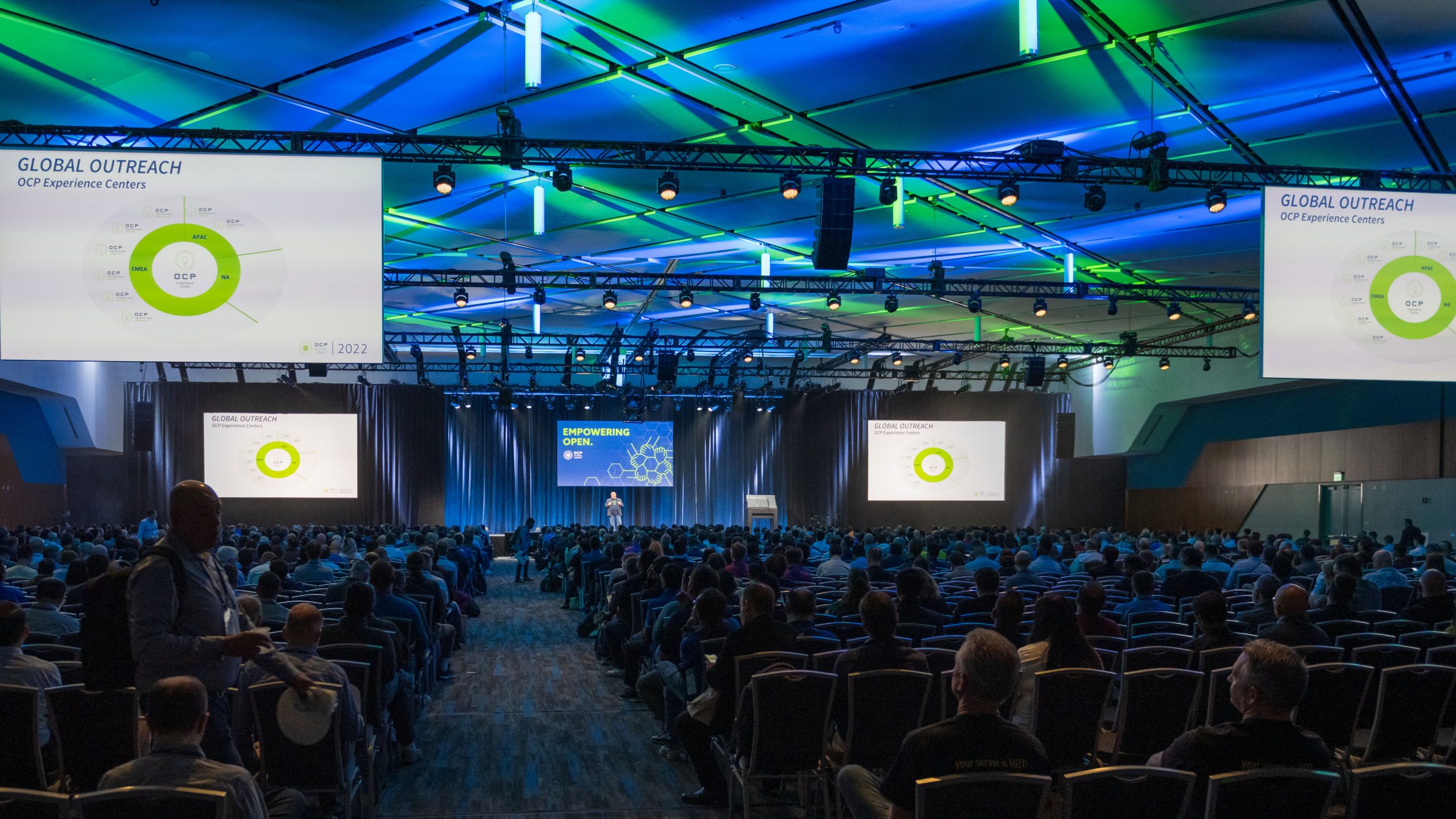 Packed general session at an OCP Summit, with large screens displaying "Global Outreach: OCP Experience Centers" as a speaker addresses the audience from the main stage